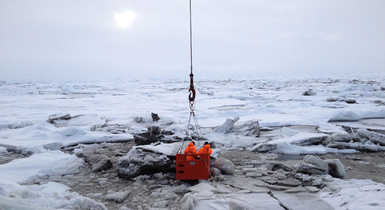 Alfred-Wegener-Institut / Rüdiger Stein | Mit unterbrochen wird auch der Transport von Sedimenten und Nährstoffen die in den flachen Zonen der arktischen Randmeere im Eis eingeschlossen werden. Das Bild zeigt von Sedimenten durchsetztes Meereis in der Transpolardrift. Zwei Forscher lassen sich per Kran von dem Eisbrecher Polarstern auf dem Eis absetzen, um Probenmaterial zu sammeln.